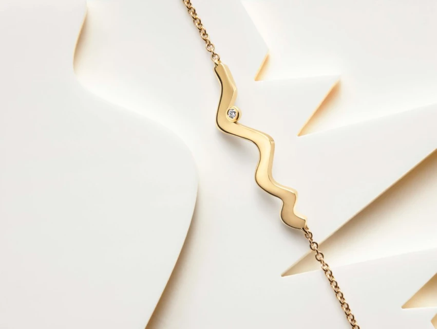 Gold necklace with a delicate chain and a wave-shaped pendant featuring a small diamond, set against a clean, bright paper surface.