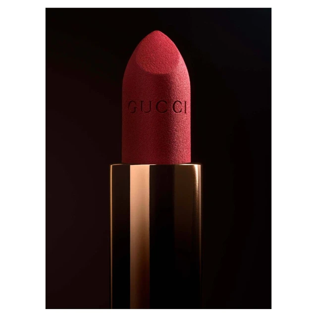 Macro shot of red Gucci lipstick featuring the logo in gold casing against a blurred black background. Beauty and fashion product shot.
