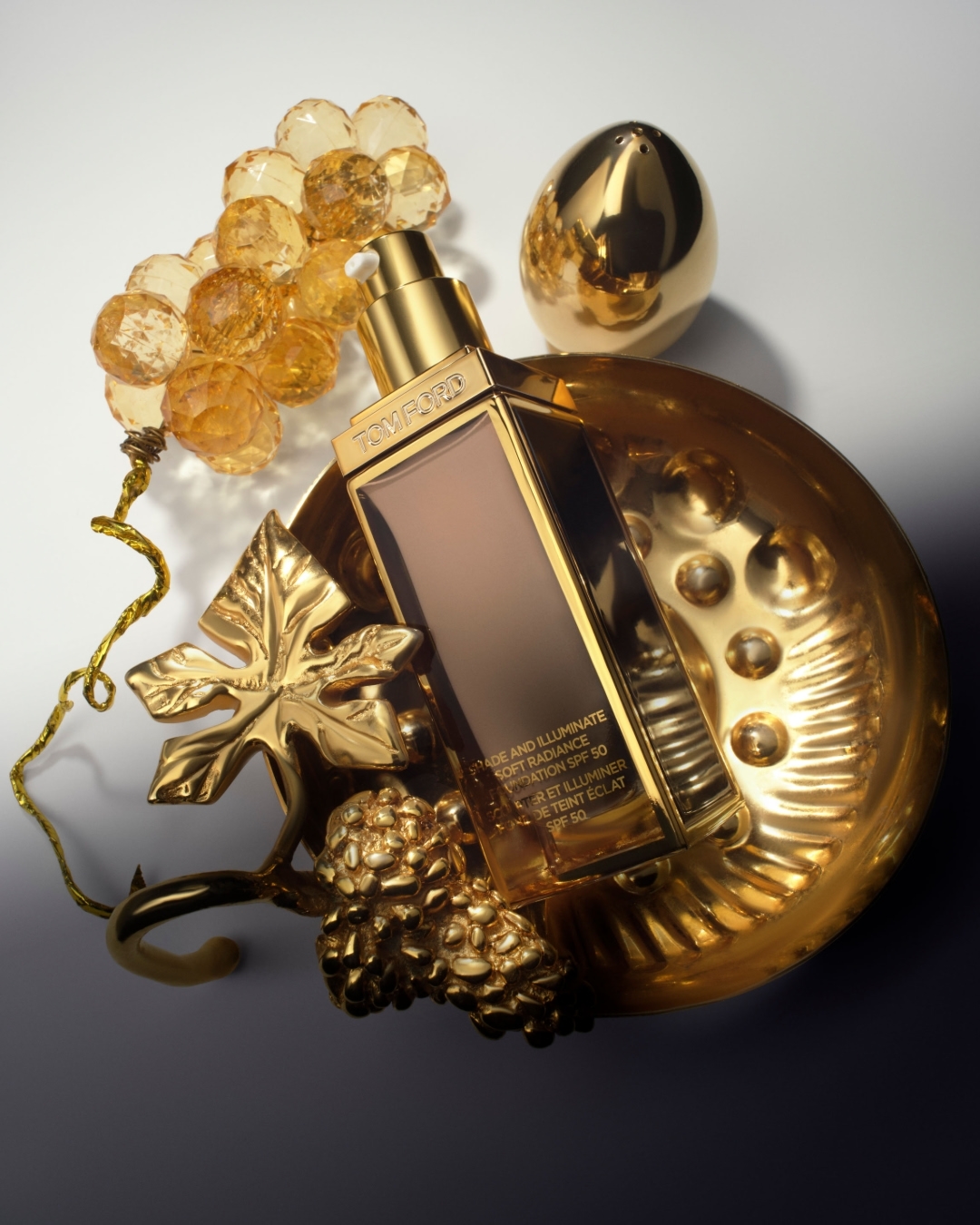 Beauty product still life featuring a gold-toned foundation bottle surrounded by decorative golden grapes, leaves, and a decorative egg.
