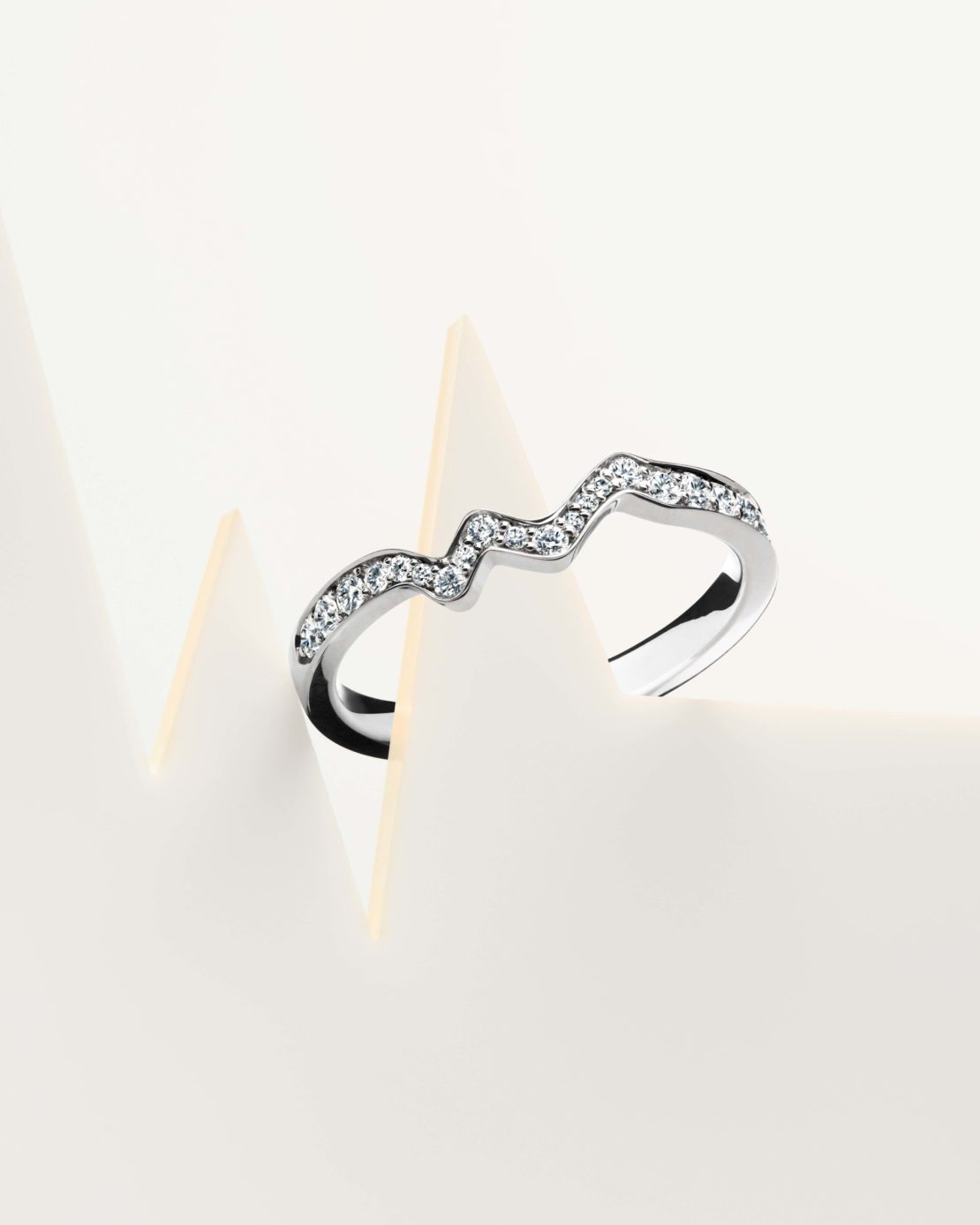 Close-up of a curved silver ring with rhinestones, displayed on a white surface with geometric paper forms, emphasizing jewelry and design.