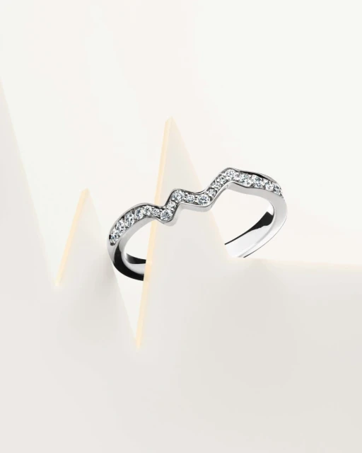 Close-up of a curved silver ring with rhinestones, displayed on a white surface with geometric paper forms, emphasizing jewelry and design.