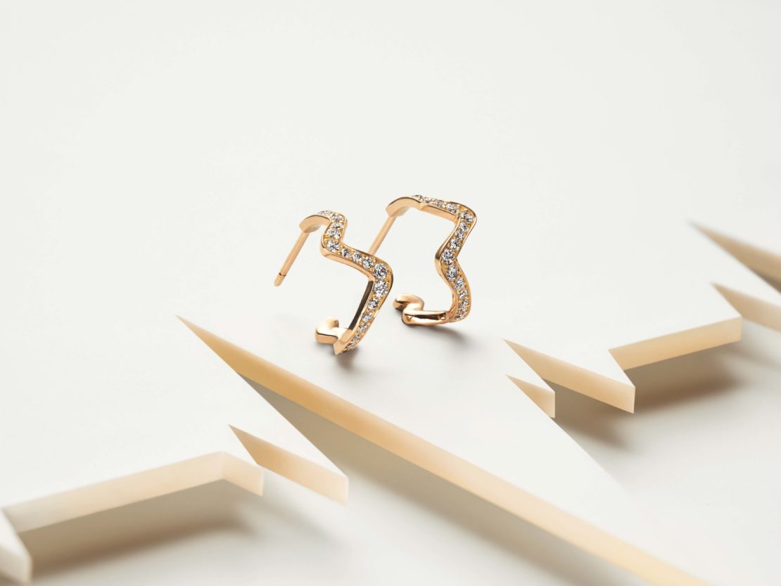 Two gold earrings with diamond accents on a white platform, showcasing jewelry design, fine metalwork, and geometric shapes in a clean studio setting.