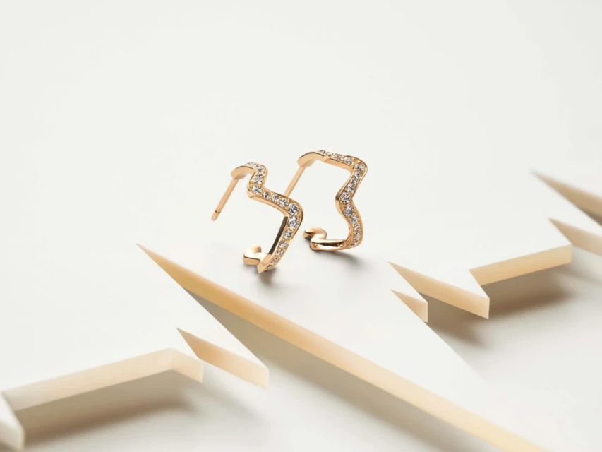 Two gold earrings with diamond accents on a white platform, showcasing jewelry design, fine metalwork, and geometric shapes in a clean studio setting.