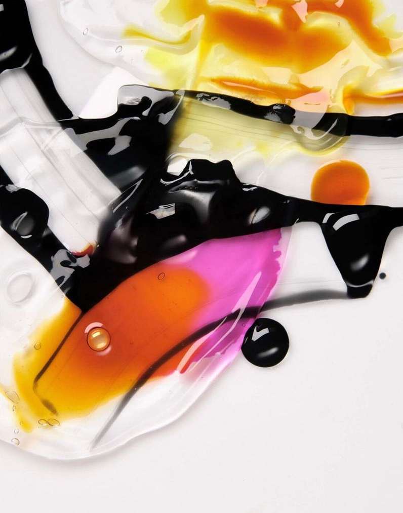 Abstract close-up of irregularly shaped liquid blobs in black, orange, pink, and yellow on a white background with a glossy finish.