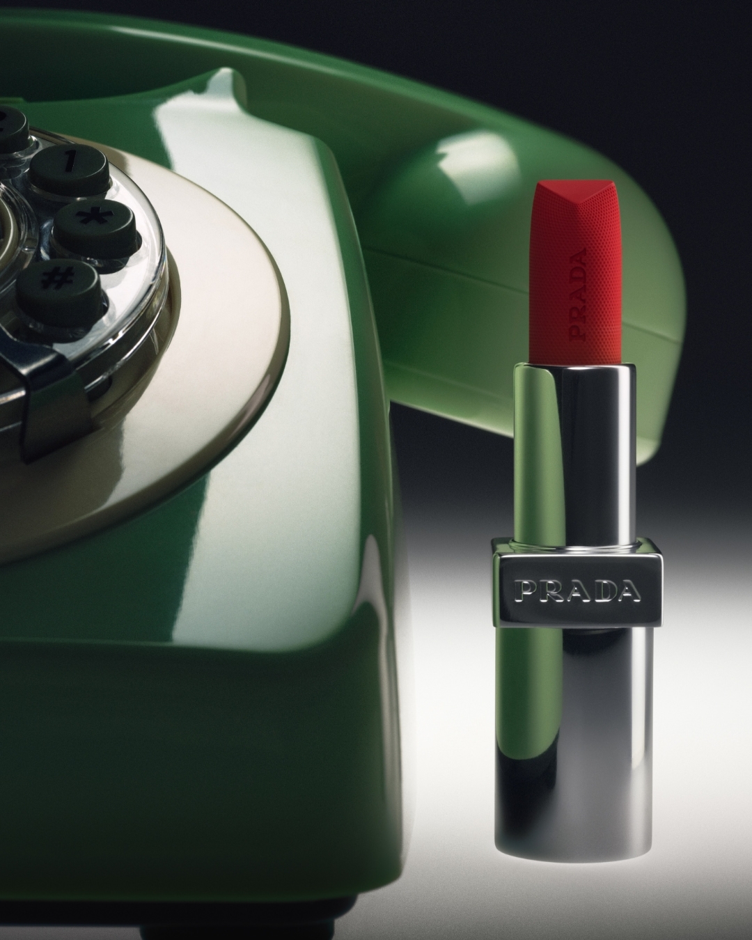 Red Prada lipstick standing upright beside a glossy green vintage rotary telephone against a gradient background