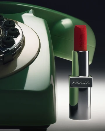 Red Prada lipstick standing upright beside a glossy green vintage rotary telephone against a gradient background