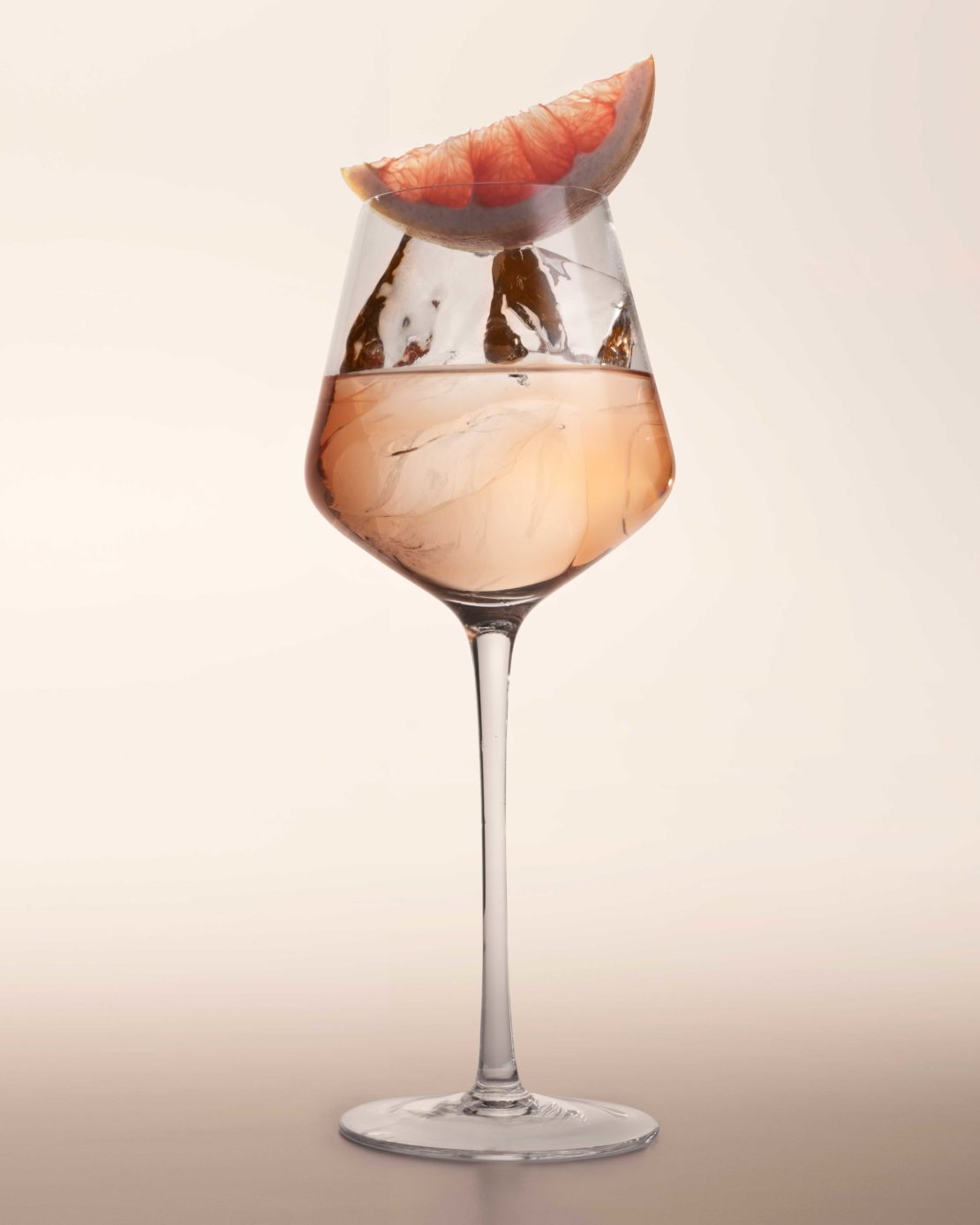 Tall glass of pink grapefruit cocktail drink with ice, isolated on light surface. Studio shot with neutral background.