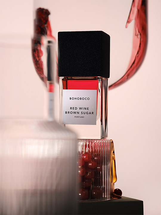 BOHOBOCO Red Wine Brown Sugar perfume bottle displayed in an artistic composition with glass containers and red liquid details.
