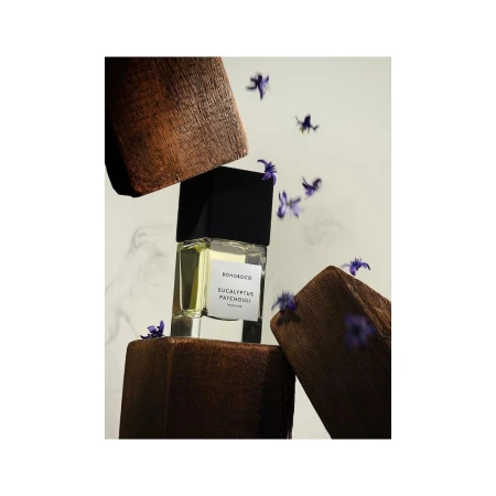 A rectangular perfume bottle surrounded by dark wooden blocks and small falling purple flowers against a light background.