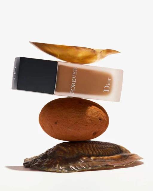 Studio shot of Dior Forever foundation bottle balanced on a rock and shells, set against a bright white background for beauty advertising.