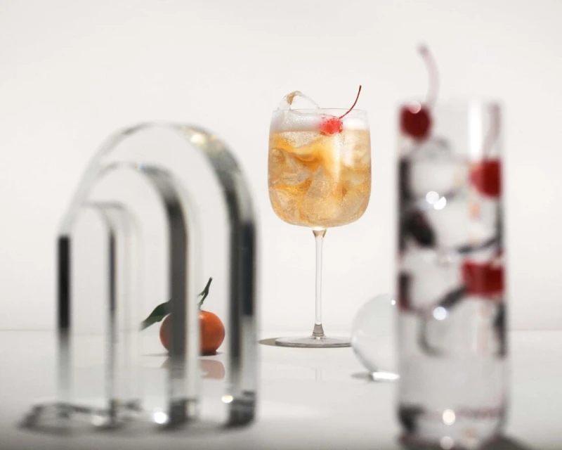 Two cocktail glasses, one with orange liquid and a cherry garnish, and another blurred glass with cherries on a white surface.