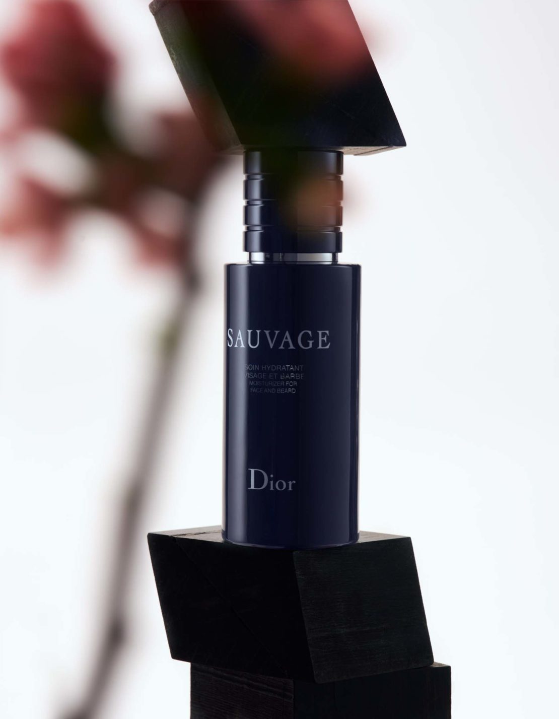 Sauvage Dior men's moisturizer bottle, navy, on black blocks, shot in a photo studio with a blurry floral background and bright, soft lighting.
