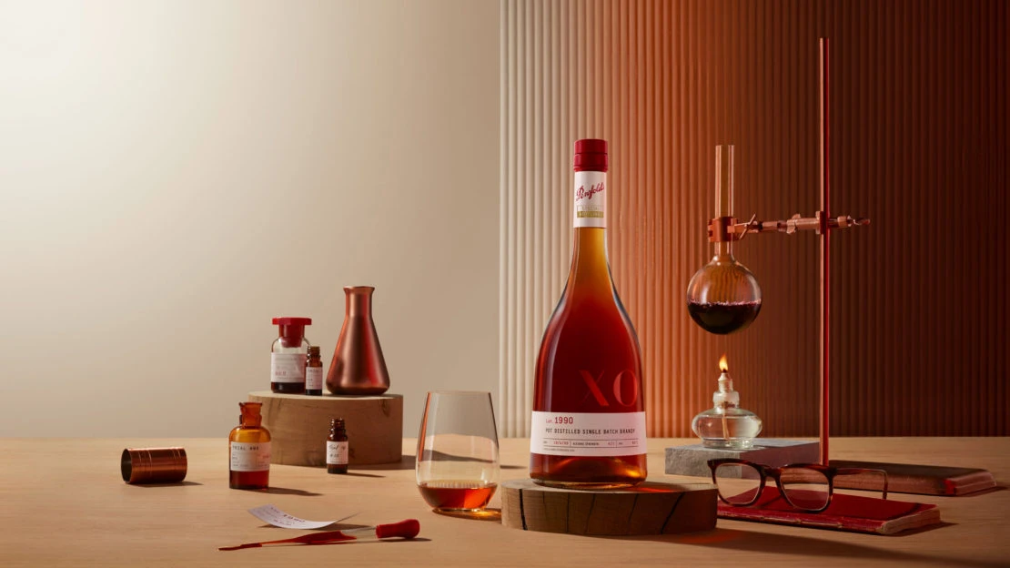 Bottle of Penfolds brandy with laboratory glassware, glasses, and books, arranged on a wooden table against a two-toned backdrop.