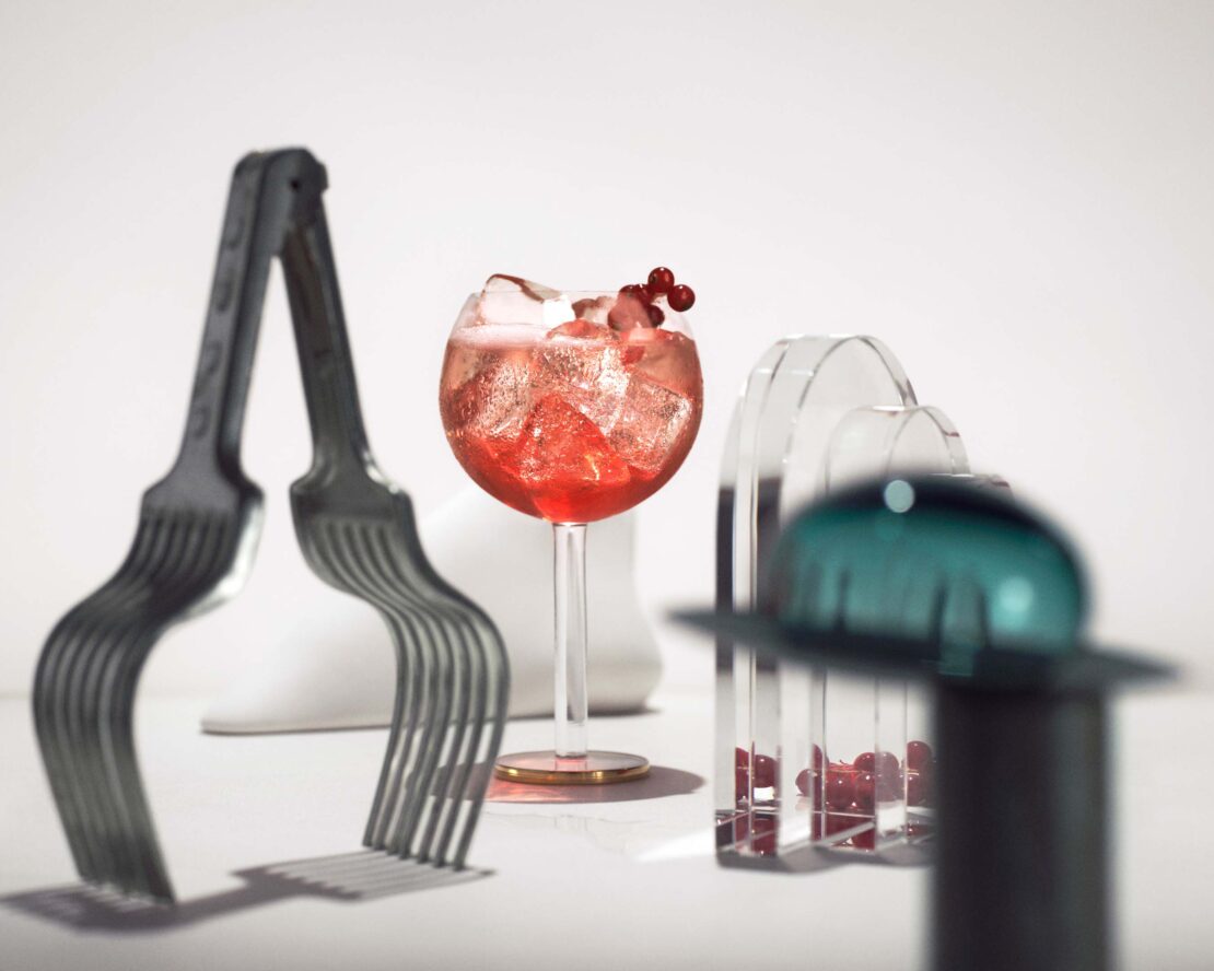 A still life photograph of a red cocktail with ice and berries, arranged with clear sculptural elements and metal fork art on a white surface.