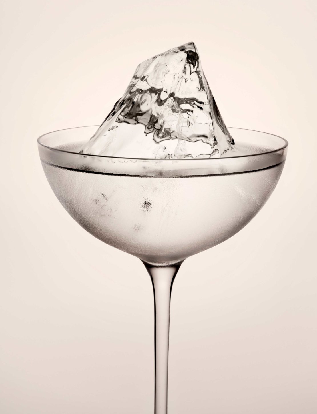 A close-up studio shot of a coupe glass filled with a clear liquid and a large ice cube, set against a soft white backdrop.