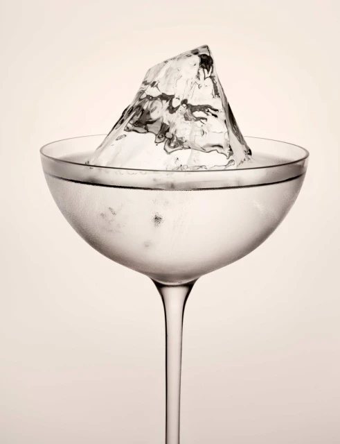 A close-up studio shot of a coupe glass filled with a clear liquid and a large ice cube, set against a soft white backdrop.