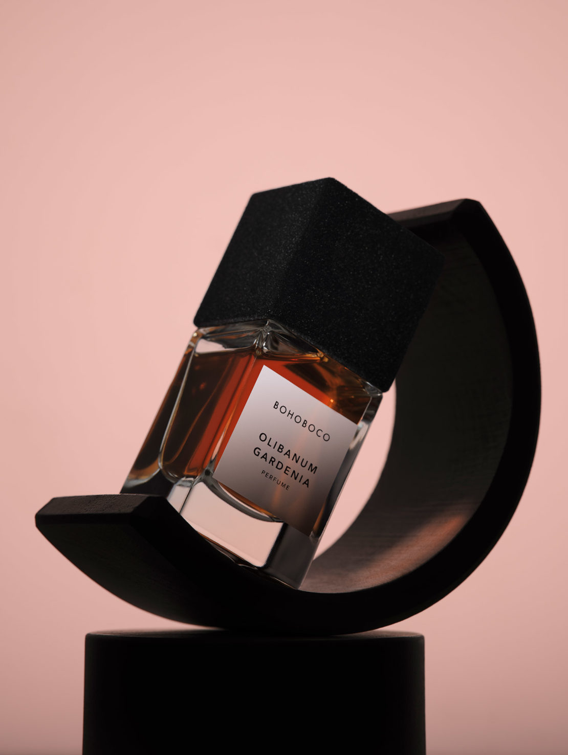 Still life studio shot of a glass perfume bottle with a black geometric cap, presented on a black sculptural stand against a pink background.