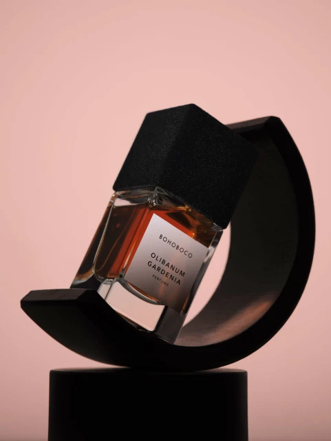 Still life studio shot of a glass perfume bottle with a black geometric cap, presented on a black sculptural stand against a pink background.