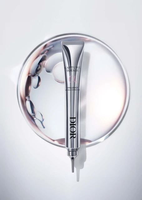 Elevated view of a Dior Capture Totale Hyalushot tube on a silver plate with swirling liquid, captured in a studio setting, showcasing beauty product.