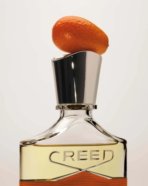 Still life of Creed Aventus cologne bottle with chrome cap, topped with a bright orange kumquat fruit on a clean white backdrop.