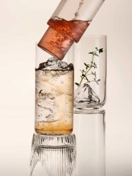 Close-up commercial shot of clear glassware with ice cubes, amber liquid, and a fresh herb sprig by Sam Hofman
