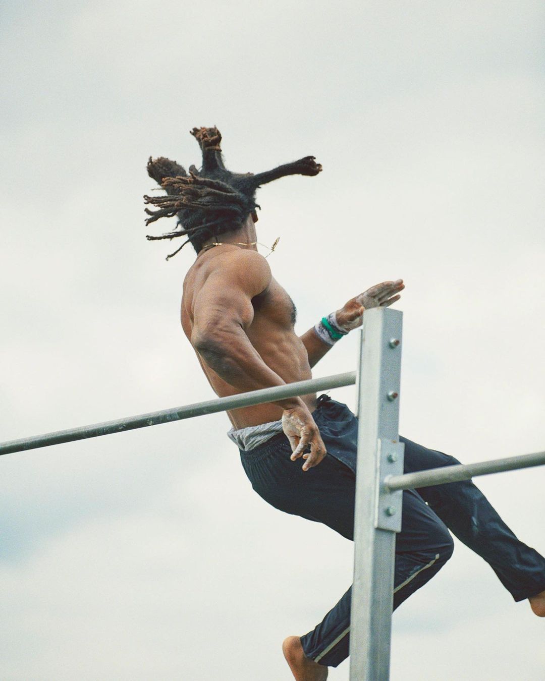 A shirtless athlete with dreadlocks performs an aerial workout on a metal bar structure against a cloudy sky backdrop, showcasing strength.