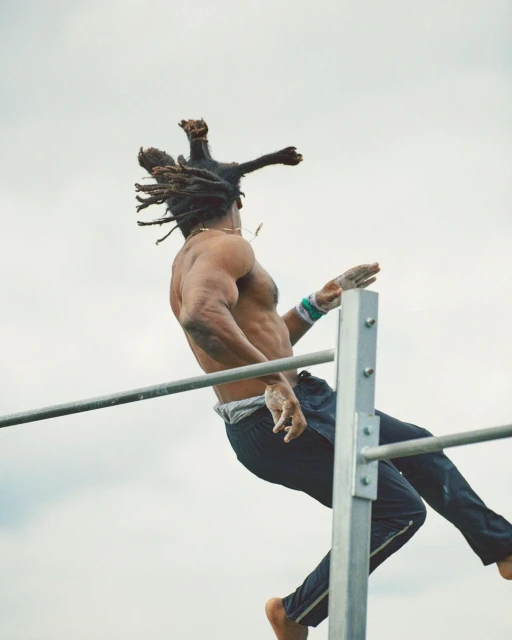 A shirtless athlete with dreadlocks performs an aerial workout on a metal bar structure against a cloudy sky backdrop, showcasing strength.