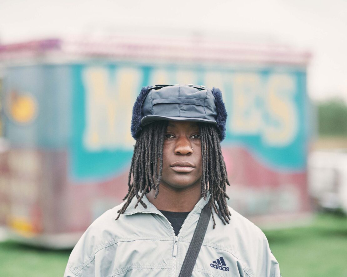 Portrait of a young adult with dreadlocks wearing a fur-lined hat and Adidas jacket outdoors against a blurred background.