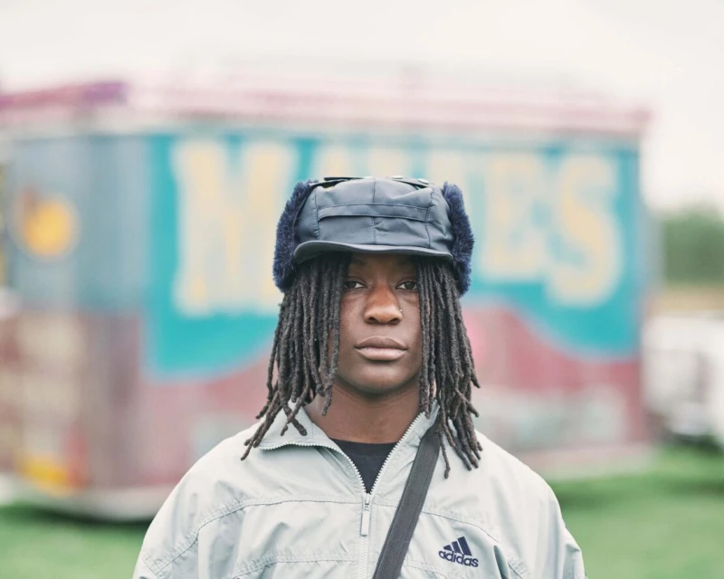 Portrait of a young adult with dreadlocks wearing a fur-lined hat and Adidas jacket outdoors against a blurred background.