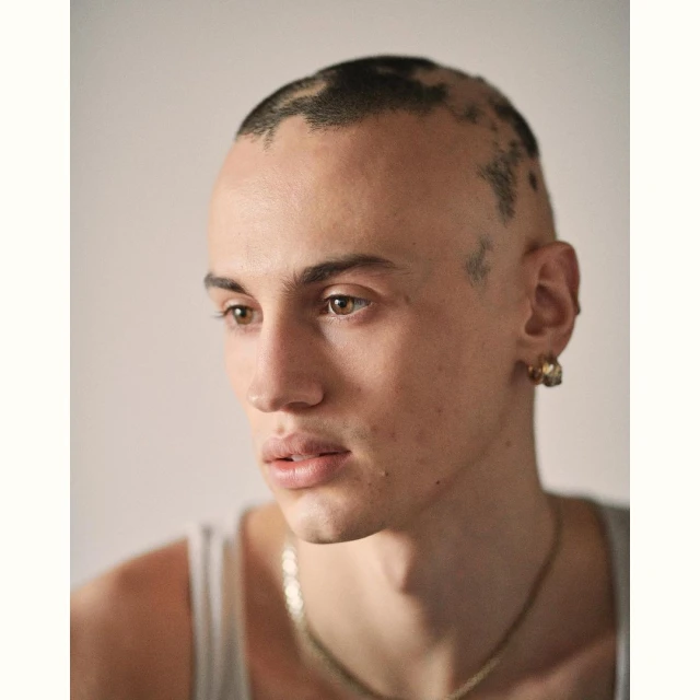 Portrait of a young adult male with a partially shaved head and gold jewelry, posing in front of a neutral background in a close-up shot.