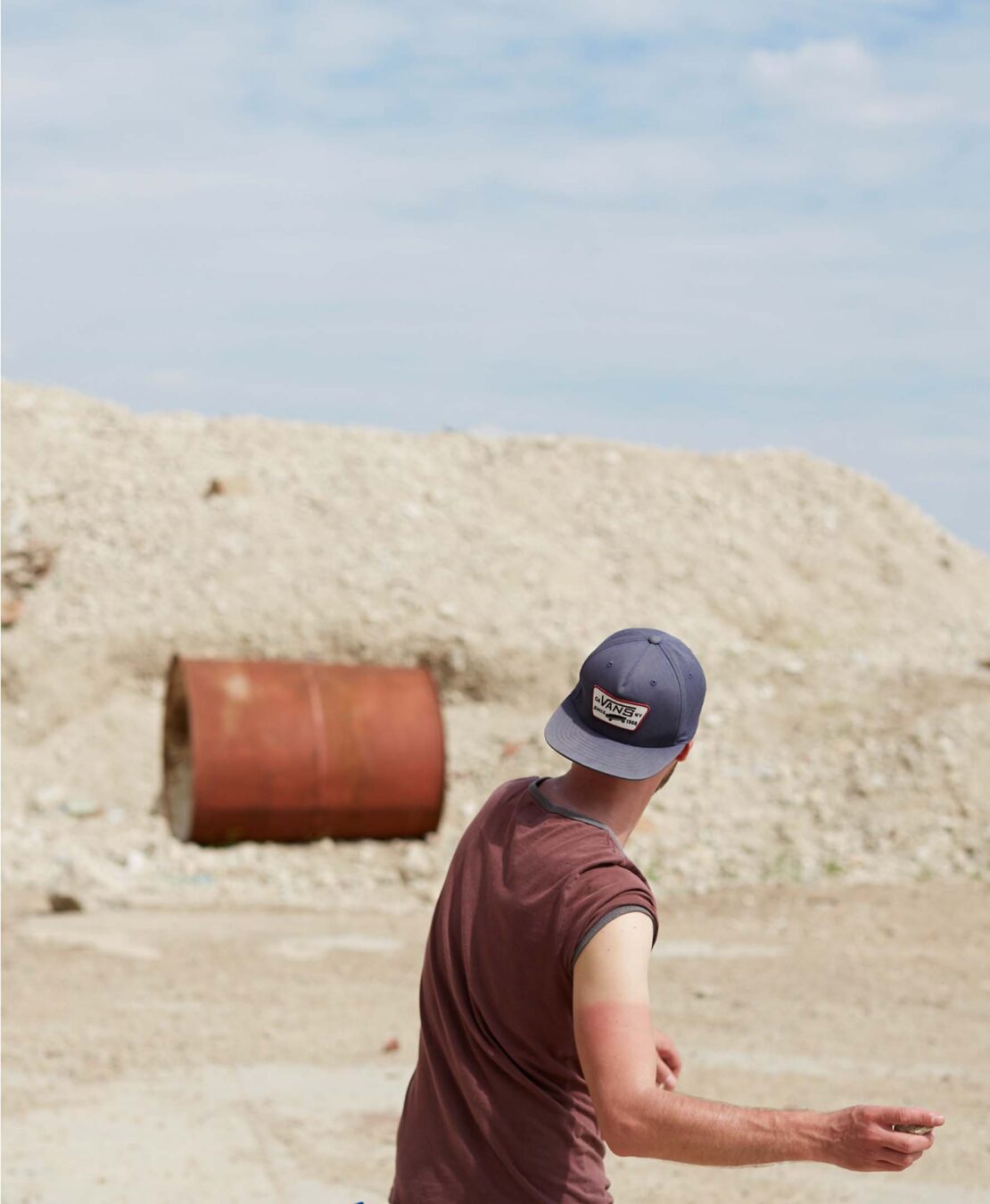 Back view of a man in a ball cap standing in a sandy outdoor environment with a rusted metal barrel in the midground.