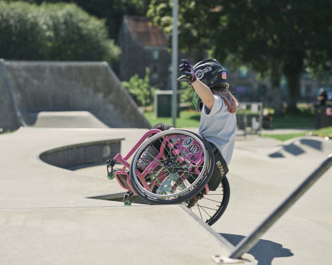 A person in a wheelchair on a ramp at a skate park raises their arms, with concrete ramps and trees in the background on a sunny day.