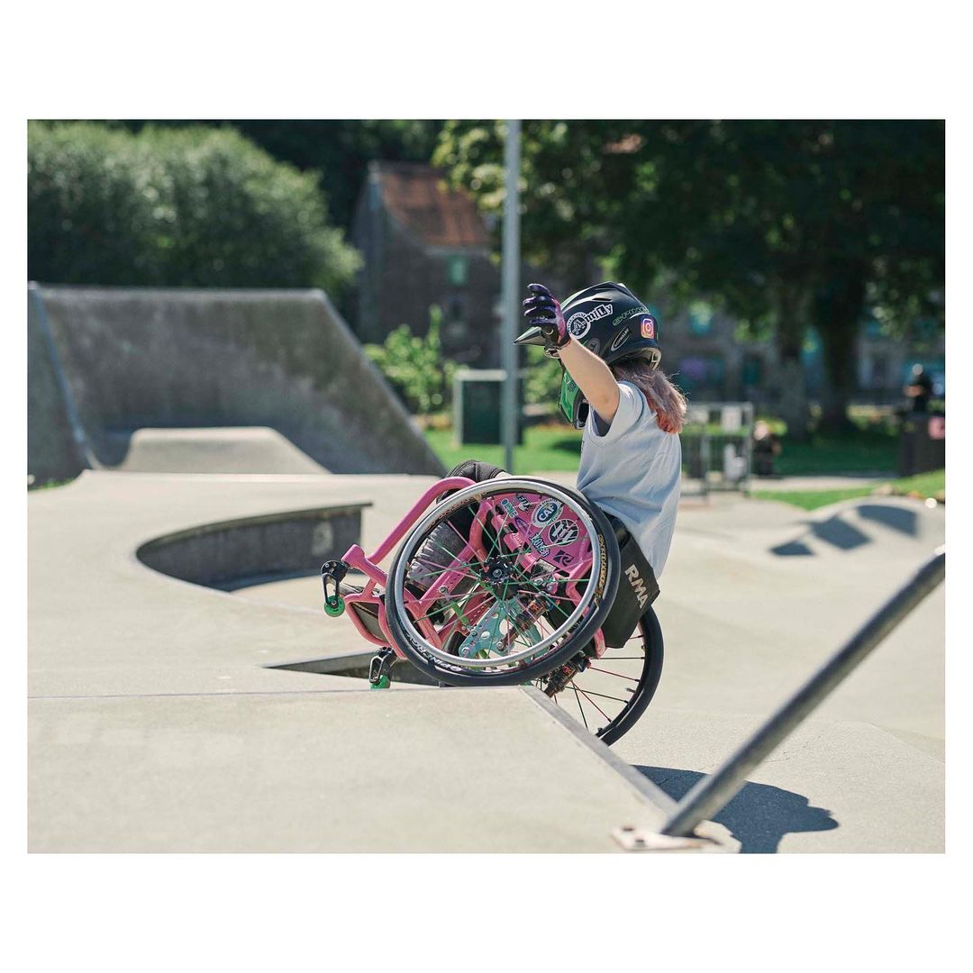A paraplegic woman wearing a helmet, gloves, and grey t-shirt balances on her pink wheelchair on the edge of a concrete ramp in a skate park.