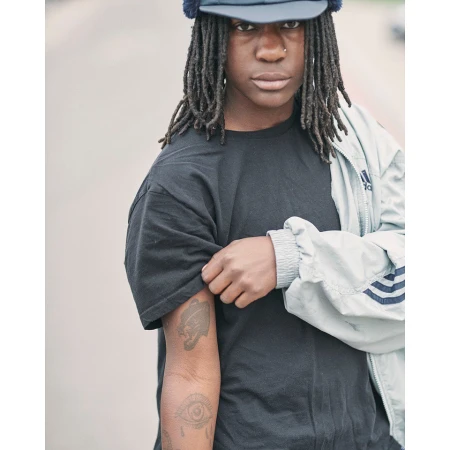 Portrait of a Black man with long dreadlocks wearing a black t-shirt, cap, and light grey jacket in London