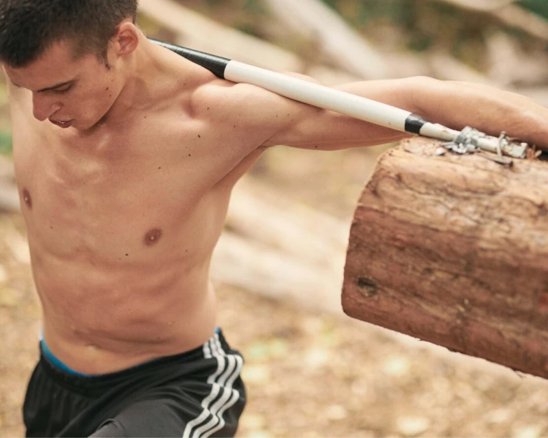 An adult male is seen from the waist up outdoors, carrying a log over his shoulders. He has a muscular build and is shirtless.