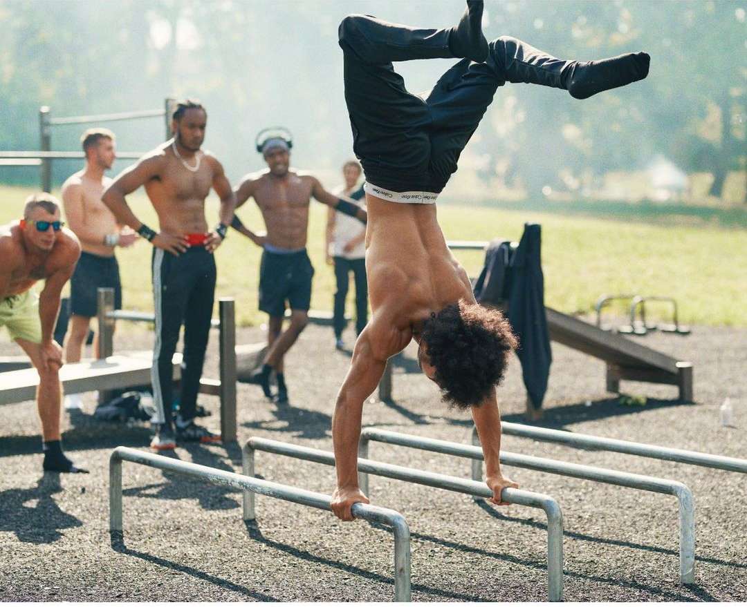 A shirtless man with curly hair performs a handstand on parallel bars outdoors while a group of men watch in the background.