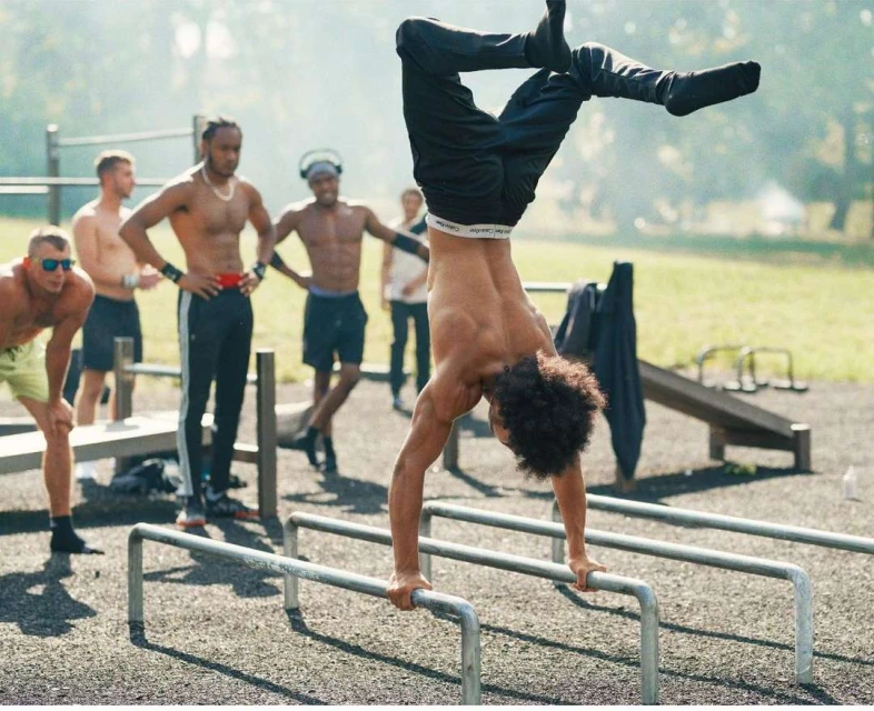 A shirtless man with curly hair performs a handstand on parallel bars outdoors while a group of men watch in the background.