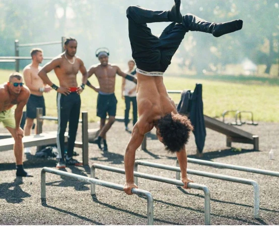 A shirtless man with curly hair performs a handstand on parallel bars outdoors while a group of men watch in the background.