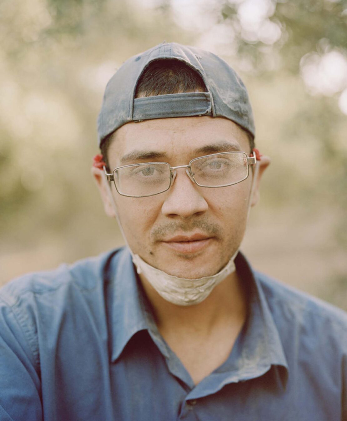 Portrait of an adult man wearing glasses and a cap, standing outdoors with a blurred background in soft, natural lighting.