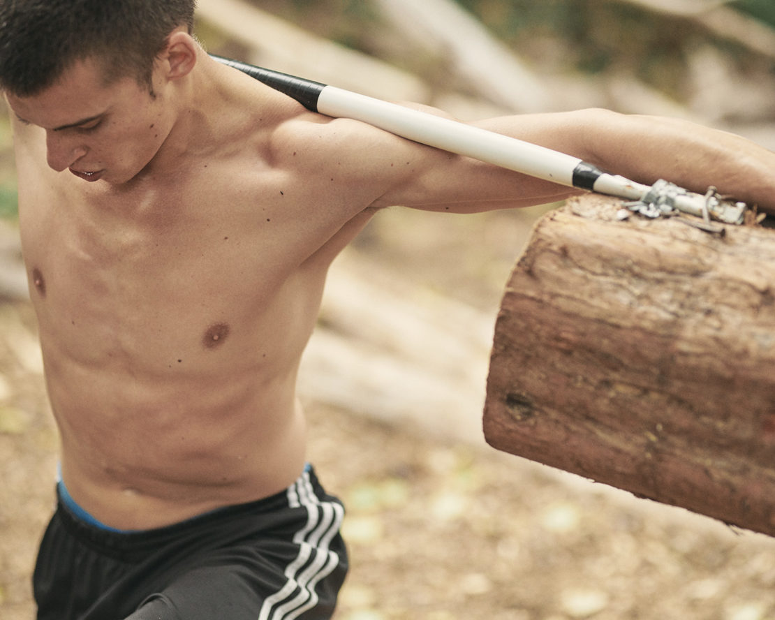 A shirtless man with a muscular build carries a wooden log over his shoulders while exercising outdoors in a natural setting.