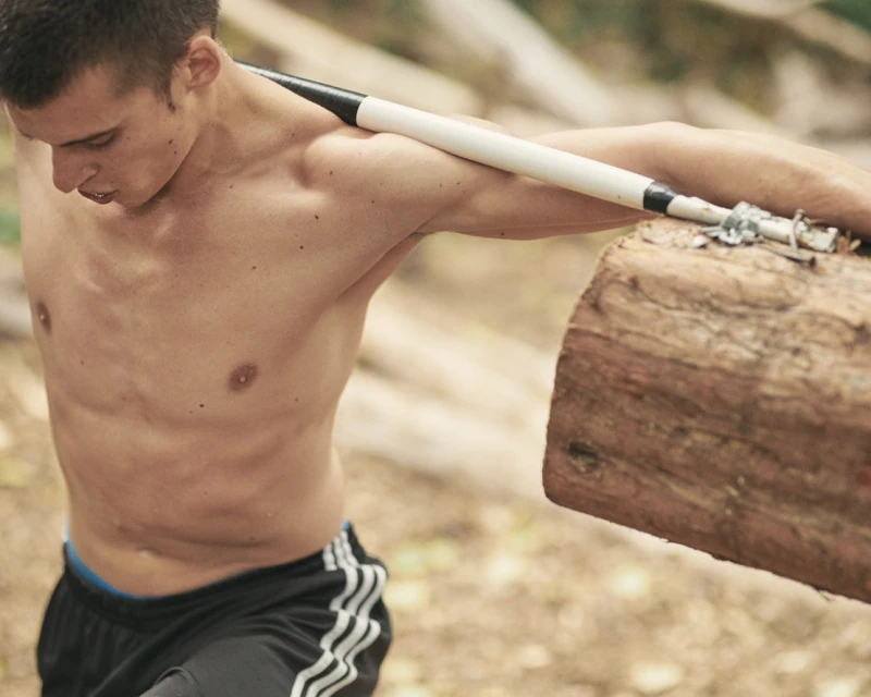 A shirtless man with a muscular build carries a wooden log over his shoulders while exercising outdoors in a natural setting.
