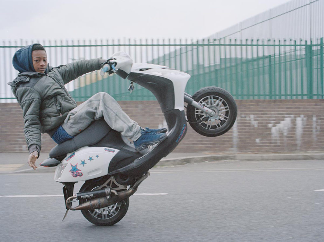 Teenage boy performs a wheelie on a white scooter in an urban setting, wearing a jacket and jeans, showcasing youthful energy and skill.