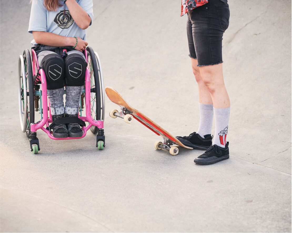 Young person in wheelchair next to a skateboard, standing near an adult on concrete, outdoors on a sunny day, skateboarding.