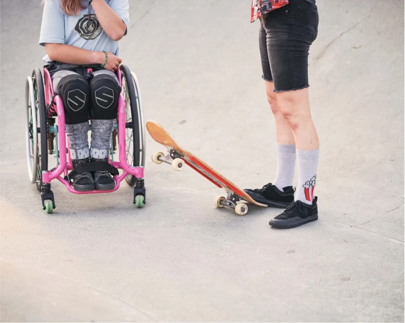 Young person in wheelchair next to a skateboard, standing near an adult on concrete, outdoors on a sunny day, skateboarding.