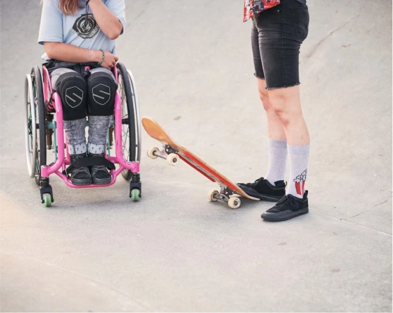 Young person in wheelchair next to a skateboard, standing near an adult on concrete, outdoors on a sunny day, skateboarding.