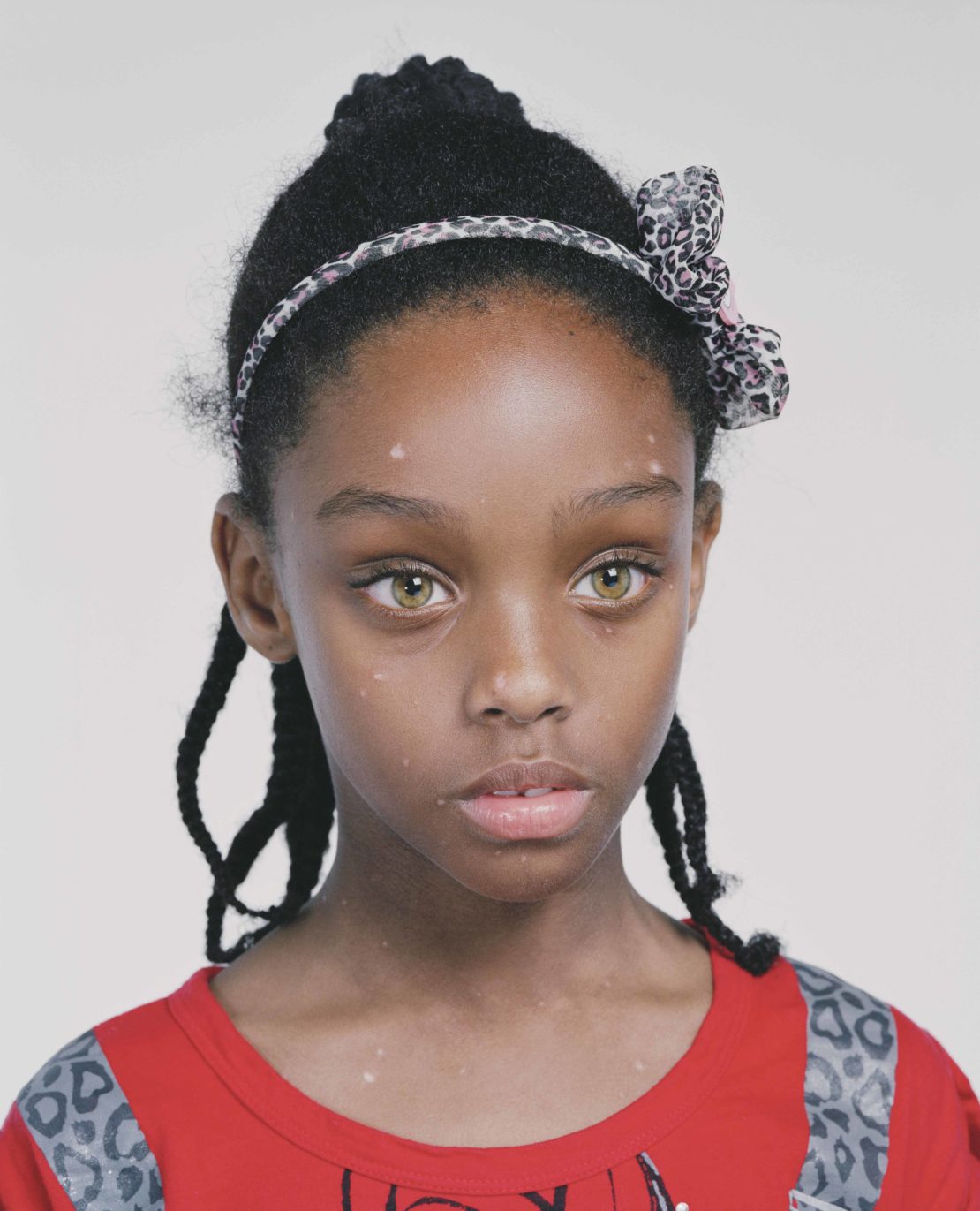 Close-up portrait of a young African-American girl with braided hair and hazel eyes wearing a red top and a leopard print headband.