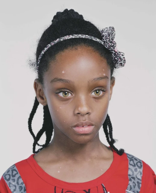 Close-up portrait of a young African-American girl with braided hair and hazel eyes wearing a red top and a leopard print headband.