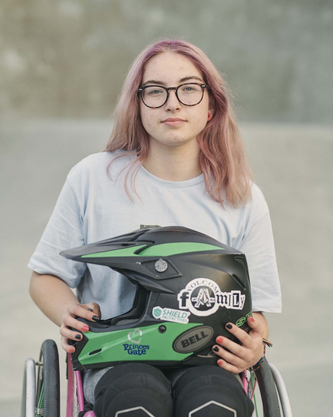 Young woman with pink hair in wheelchair, holding black and green helmet, outdoors in daylight; portrait style photo.