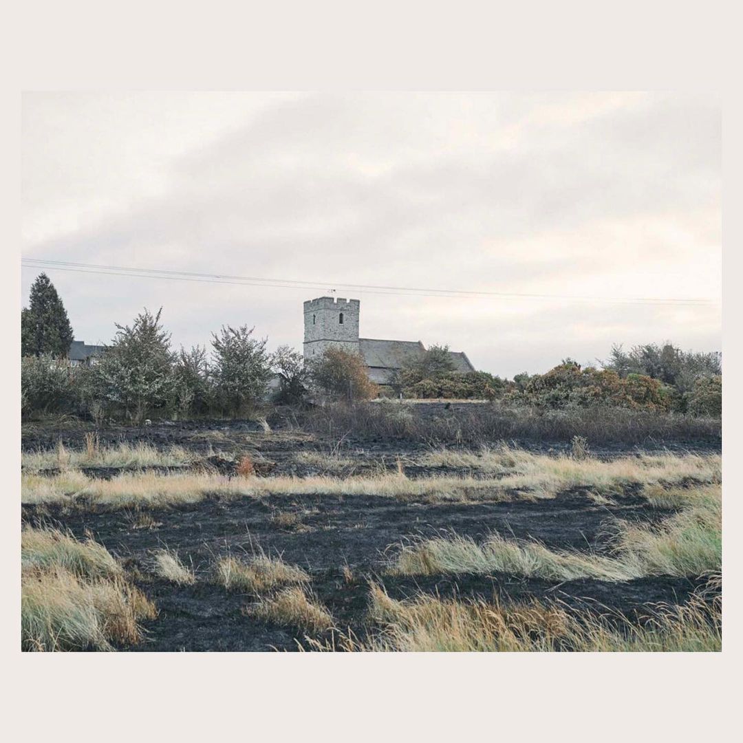 Rural landscape featuring a stone church in the background, with a field of dry grass and dark, burnt patches in the foreground.