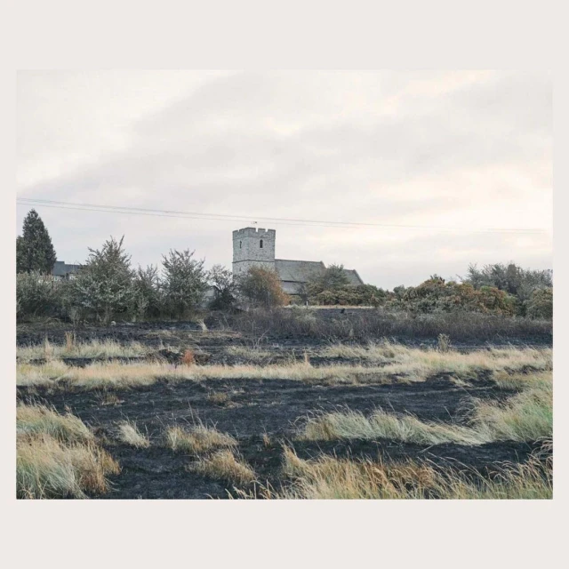 Rural landscape featuring a stone church in the background, with a field of dry grass and dark, burnt patches in the foreground.